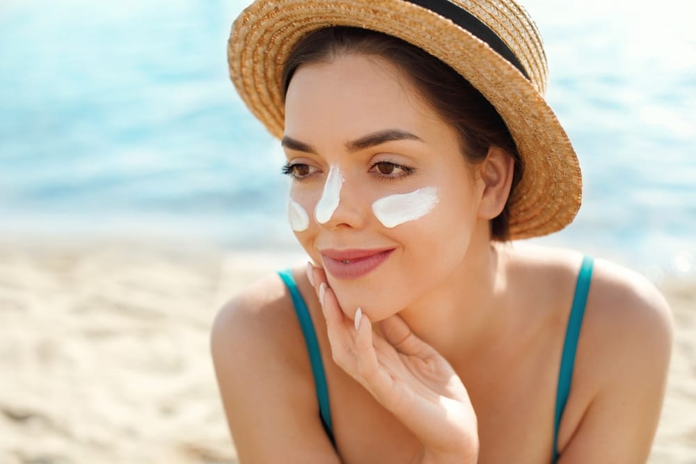Beautiful woman applying cream sunscreen on a tanned face while at beach and other traveling