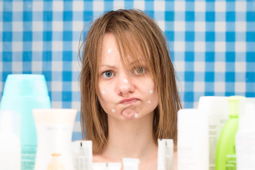 Close-up portrait of upset cute girl without makeup, with white spots of cosmetic on her problem face skin, standing in bathroom surrounded by various cosmetic products as recommended on TikTok