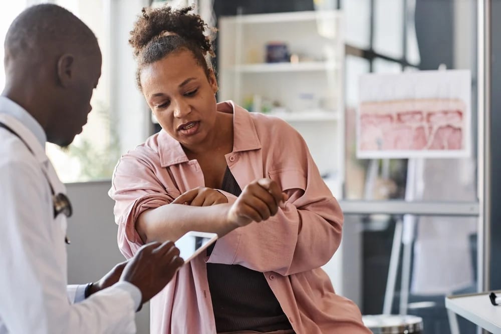 Adult African American woman suffering from psoriasis visiting dermatologist in clinic and pointing on rash spots on arms