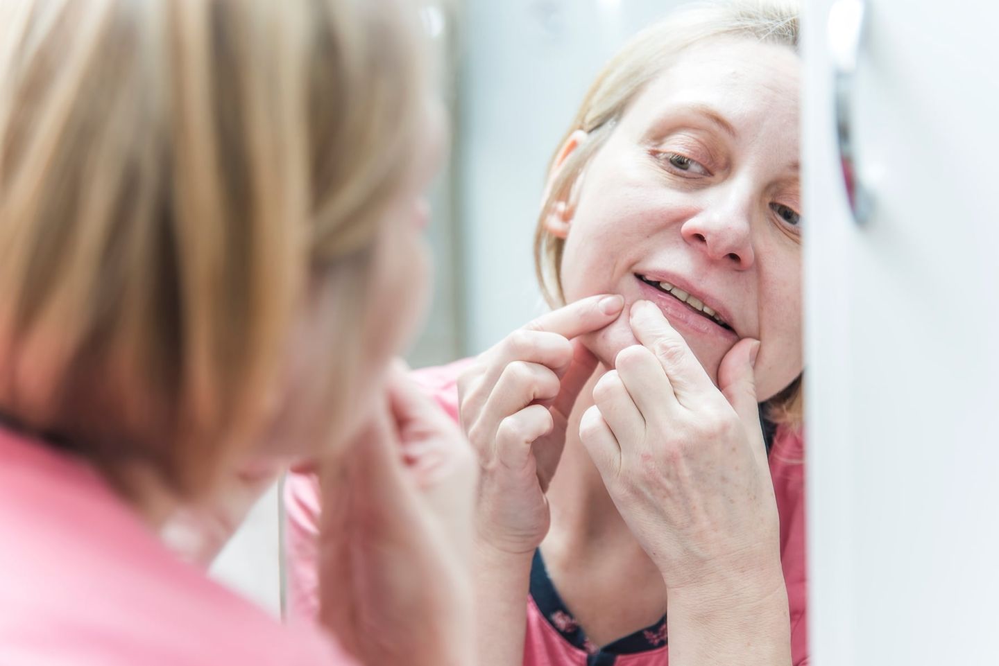Mature blonde woman in her pajamas, squeezing pimples, helping herself with a bathroom mirror