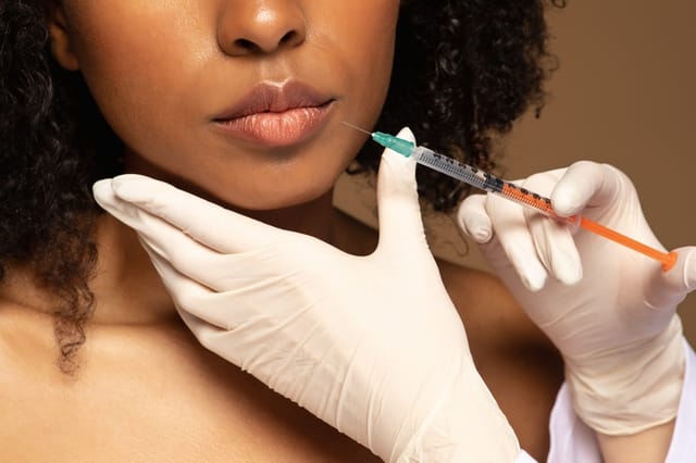 Closeup of beautician hands in white medical gloves holding syringe with beauty filler next to african woman beautiful juicy plump lips