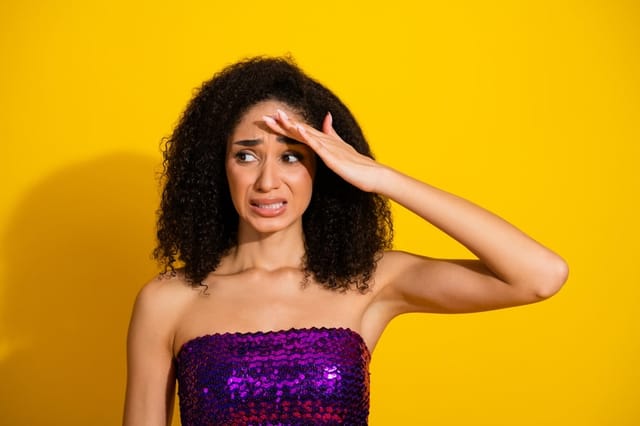 Young woman with curly hair wearing a purple sequin top shields her eyes against a bright yellow background stressed mood affecting skin health