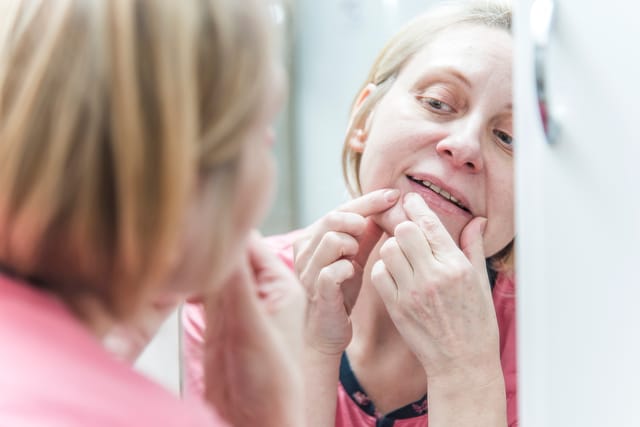 Mature blonde woman in her pajamas, squeezing pimples, helping herself with a bathroom mirror