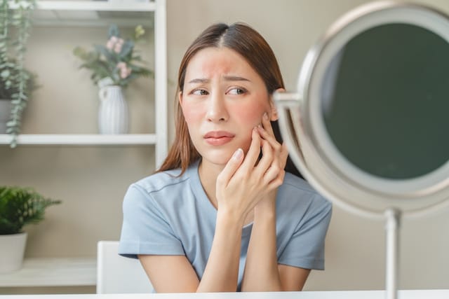 Asian young woman looking at red spot on her face from cosmetic allergy or sensitive skin overtreated in the mirror