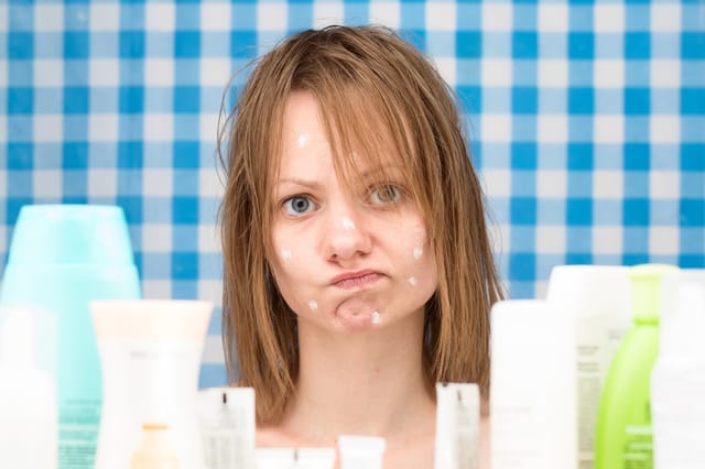 Close-up portrait of upset cute girl without makeup, with white spots of cosmetic on her problem face skin, standing in bathroom surrounded by various cosmetic products as recommended on TikTok