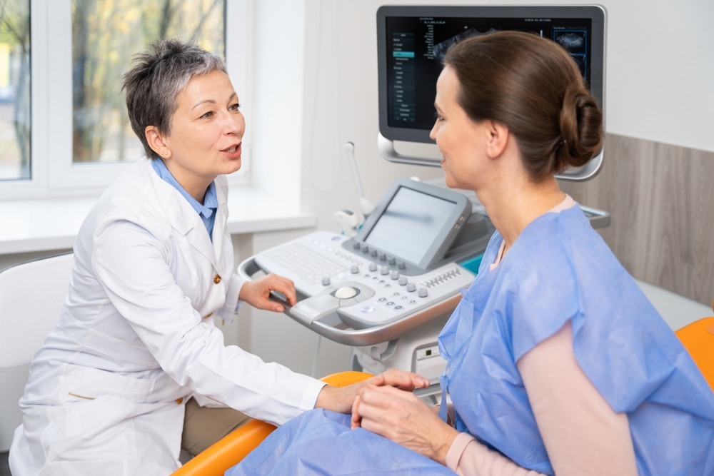 A doctor and patient converse during an ultrasound appointment, with the doctor operating the machine and the patient in a blue gown, seated comfortably